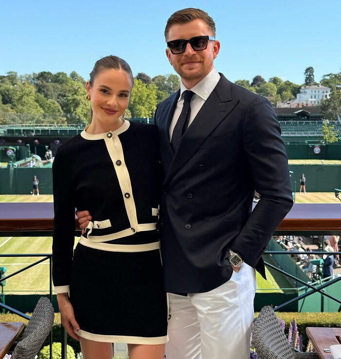 Adam Peaty posing with a woman at an outdoor sports event, dressed in formal and stylish attire.