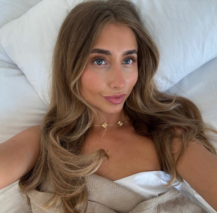 Adult star with long hair lying on a bed, wearing a delicate gold necklace, posing for a close-up selfie.