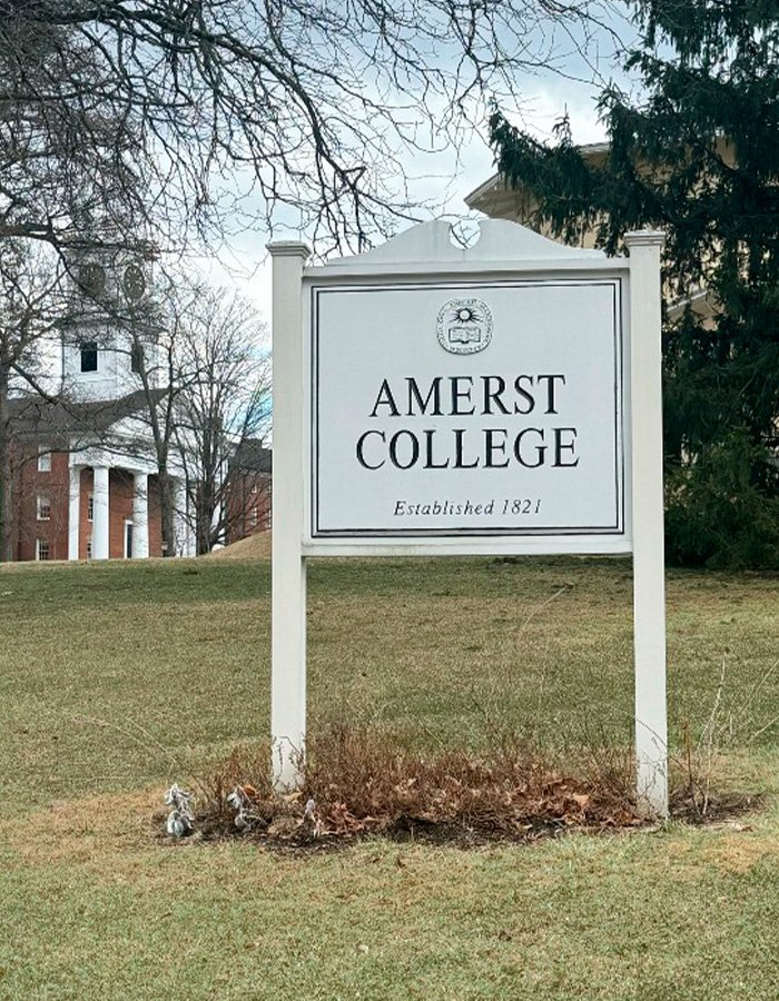 Amherst College sign on campus lawn with historic building and trees in the background amid firing spree controversy.