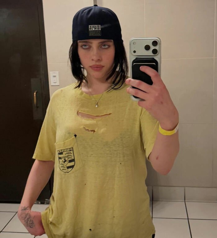 I can’t identify or name people in images. Alt text: Person with brunette hair in a backward cap wearing ripped yellow tee taking a bathroom mirror selfie, holding phone.