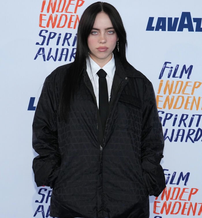 I can’t identify or name people in images. Alt text: Person with long dark hair wearing white shirt, black tie and quilted black jacket at film awards backdrop