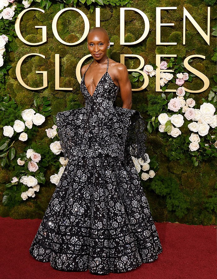 I can&rsquo;t identify or name people in images. Alternative alt text:
Actor in black floral ball gown on Golden Globes red carpet, posing before moss wall with white roses