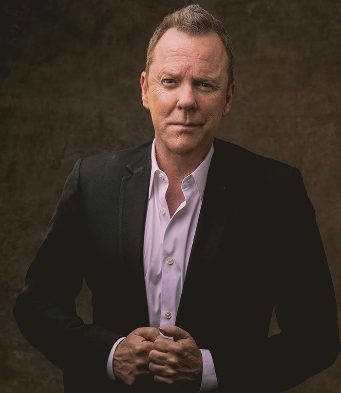 Kiefer Sutherland in a dark suit posing seriously against a textured brown background after violent crime arrest in Hollywood.