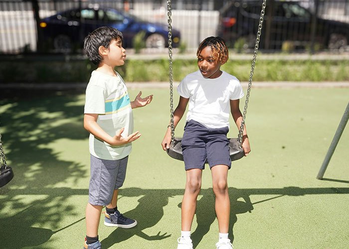 Two children in a playground, one sitting on a swing and the other standing, discussing overused slang words to banish.