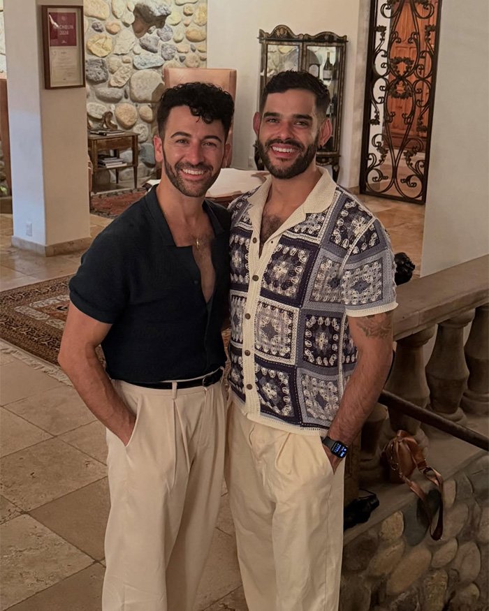 Gay couple smiling and posing indoors, one in patterned short-sleeve shirt and the other in dark open-collar top