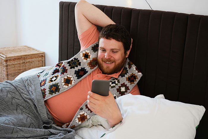 Man smiling at phone while lying in bed, emotionally invested in random family group chat conversation.