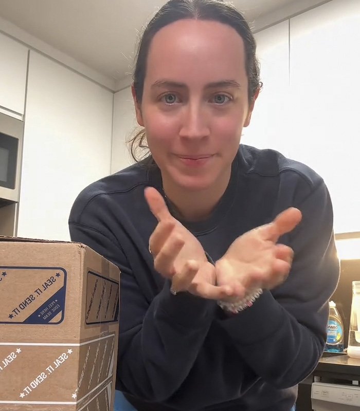 Woman in a kitchen gesturing with hands near a box, illustrating fiancé’s mug gift sparking relationship debate online.