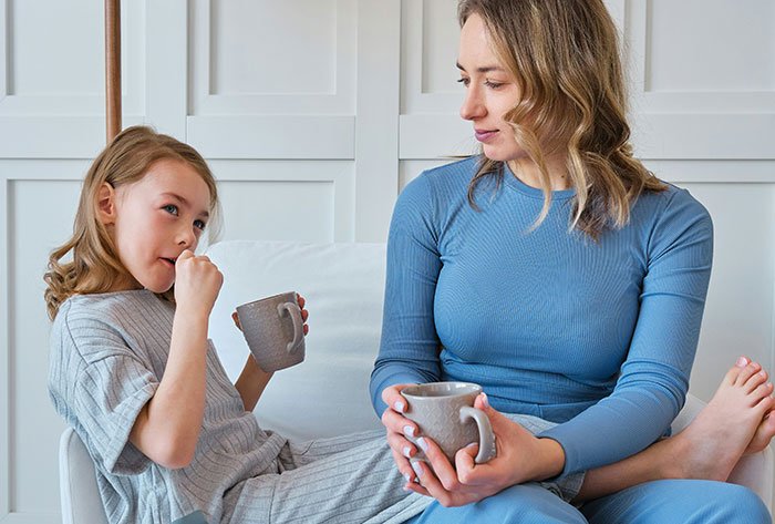 Mother and daughter on couch holding mugs, parent conflicted whether to make daughter attend birthday