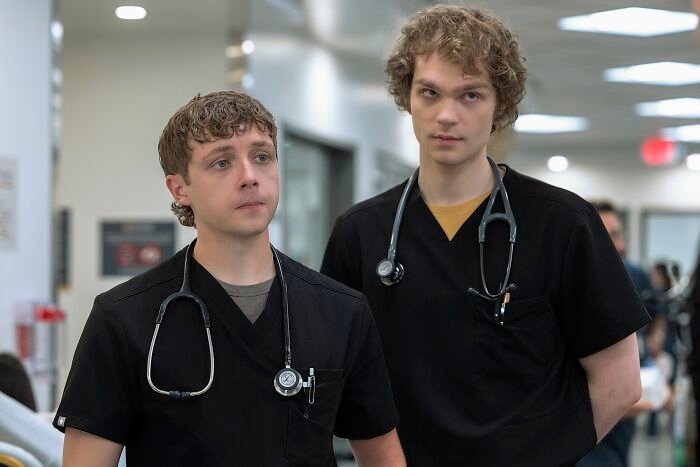 Two young male medical interns wearing black scrubs and stethoscopes in a hospital hallway, Insufferable Pitt fans discussion.