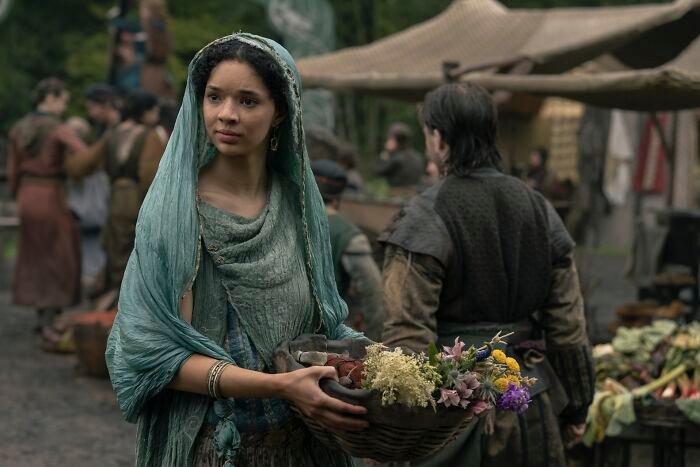 A woman in a green hooded cloak holding a basket of flowers in a crowded medieval market setting, discussing Tanzyn Crawford height.