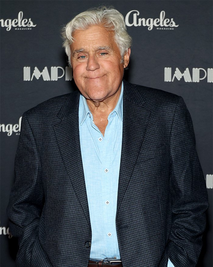 Jay Leno wearing a dark checked blazer and light blue shirt at a media event, looking thoughtful and composed.