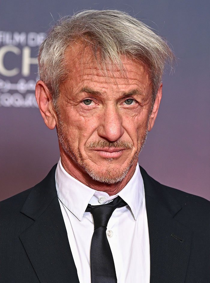 Sean Penn, 65, wearing a black suit and tie, posing at a formal event with a serious expression.
