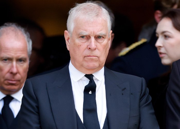 Prince Andrew in a formal black suit and tie, surrounded by people, amid controversy over emails to Epstein.