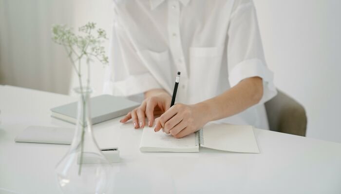Person writing in a notebook with a pencil, surrounded by books, representing smart words and well-spoken communication.