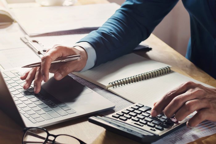 Person using a calculator and laptop at a desk with a notebook, managing finances related to emptying father's estate.