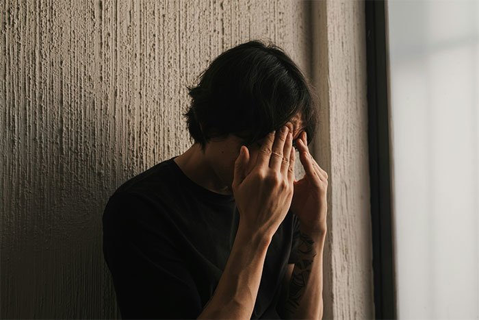 Teen boy sitting alone in a dim room, covering his face showing fear and distress related to violent sister and family issues.