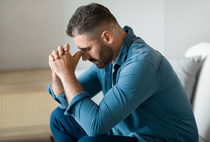 Man in a blue shirt sitting stressed with hands clasped, representing husband agreeing to an open marriage situation.