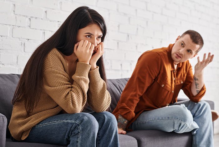 Woman upset on couch threatening to take ex’s dog to shelter while ex looks on, not intervening in the argument.