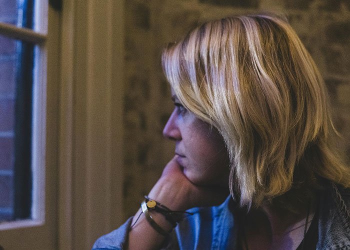 Woman feeling bad and ungrateful while reflecting on husband's abhorrent gifts by a window in a dimly lit room.