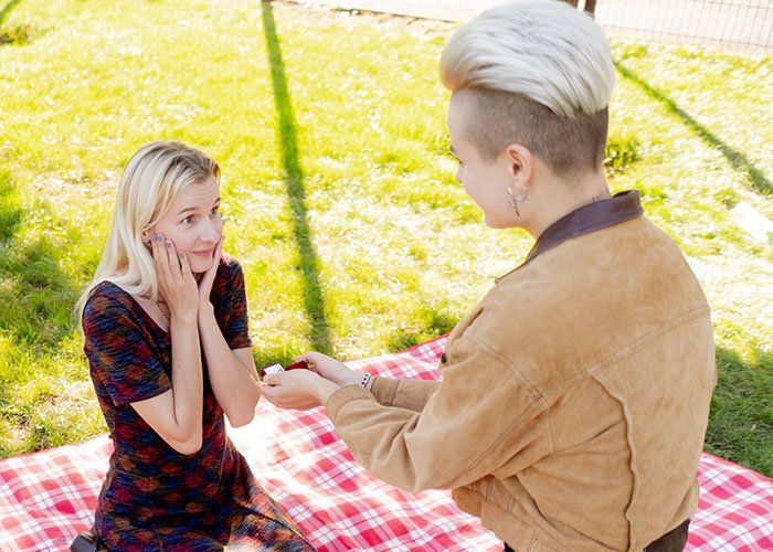 A person proposing outdoors on a red plaid blanket with a surprised fiancée showing an emotional reaction.