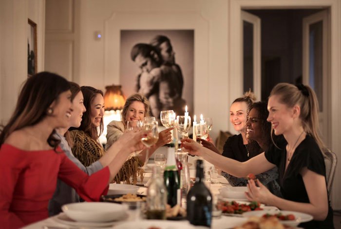 A group of women raising glasses at a dinner table, highlighting a woman refusing to pay for friends&rsquo; babysitter.