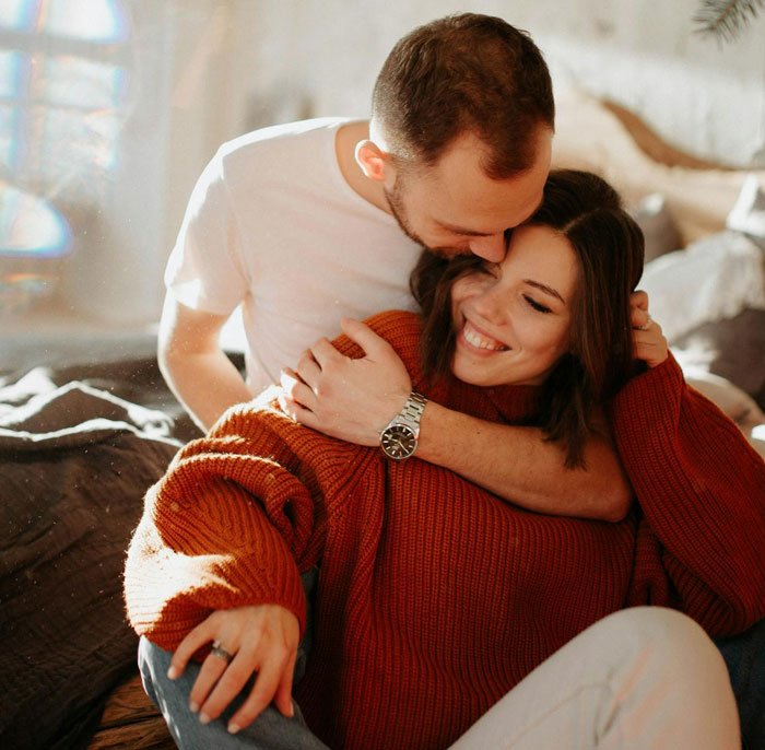 Unemployed woman embraces jobless man warmly indoors, smiling while man kisses her on the head in cozy setting.