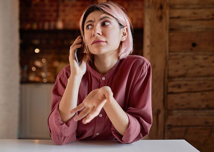 Woman demands friend to kick husband out of romantic getaway, shown frustrated while talking on phone indoors.