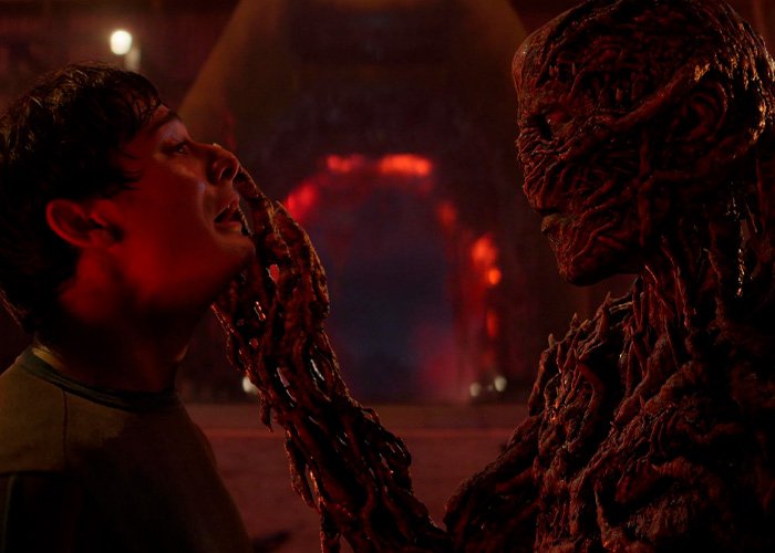 Noah Schnapp as Will Byers in Stranger Things facing a creature with a dark, twisted hand in a red-lit scene.