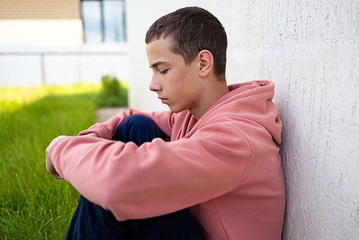 Teen sitting alone outside, looking upset and overwhelmed being a third parent to seven kids at his mom&rsquo;s house.