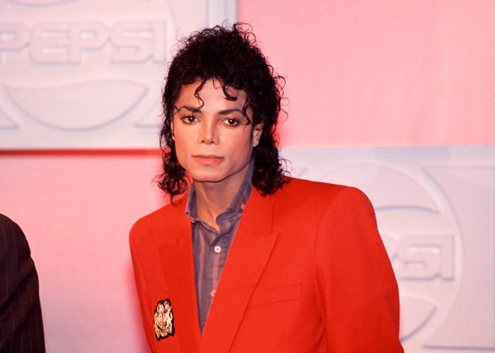 Michael Jackson wearing a red blazer with curly black hair at a promotional event, linked to secret tapes on children.