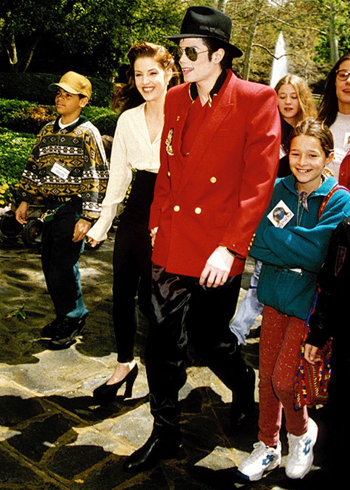 Michael Jackson walking outdoors with children and adults, wearing a red jacket and black hat in a sunny park setting.