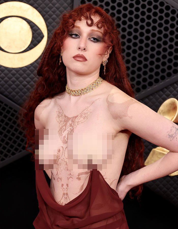 Chappell Roan posing on the Grammy red carpet with intricate body art and bold makeup in a sheer maroon outfit.