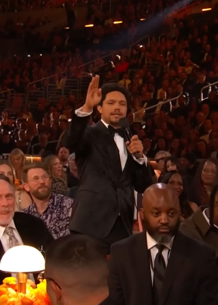 Trevor Noah in a black tuxedo speaking into a microphone at the Grammys, sparking wild theories among the audience.