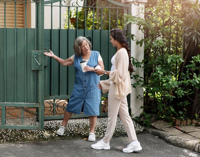 Two women talking near a green metal gate by a living hedge dividing their properties outdoors.