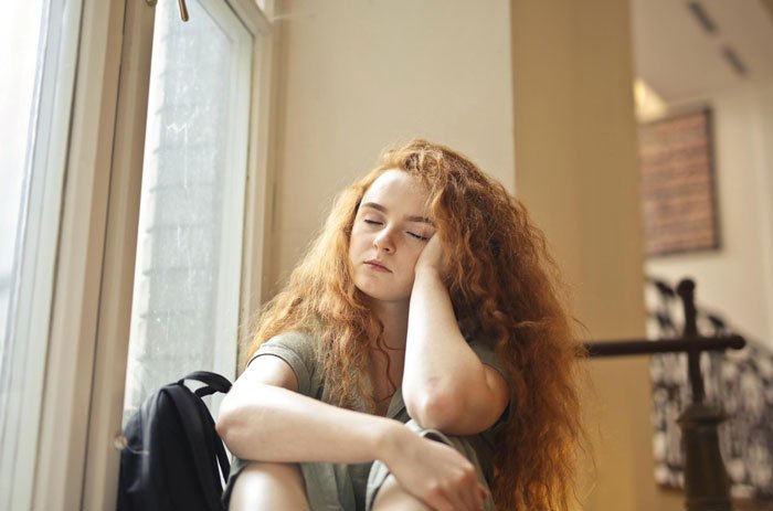 Teenage sister with red hair sitting by a window looking upset, reflecting on family and parenting drama.
