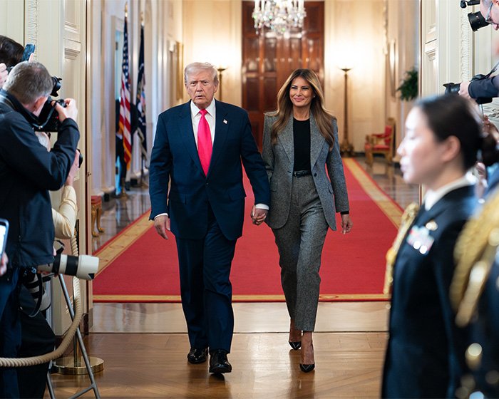 Donald Trump and Melania walking hand in hand down a hallway lined with photographers and military personnel.
