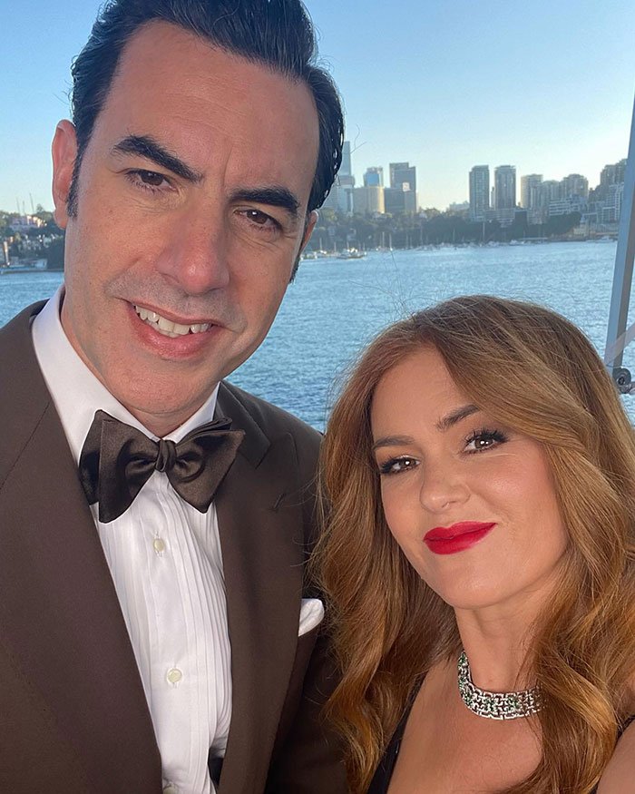 Sacha Baron Cohen and Isla Fisher posing together outdoors near water with city skyline in the background.