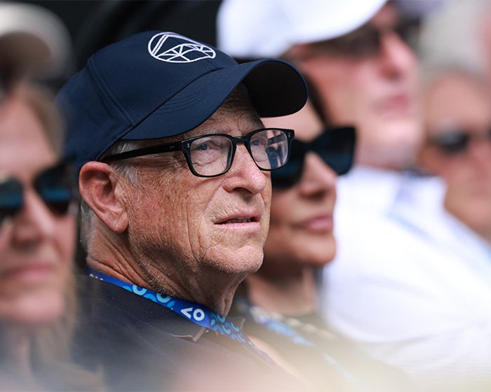 Bill Gates wearing glasses and a cap, attending an outdoor event with people blurred in the background.