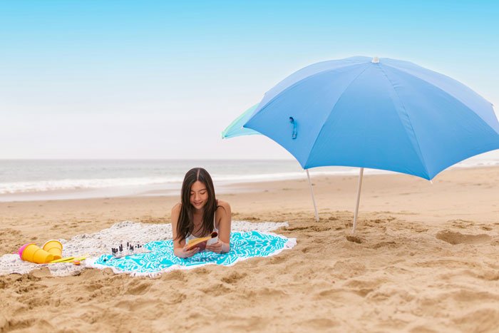 Woman reading on beach under large blue umbrella, illustrating woman makes husband post argument online reality check.