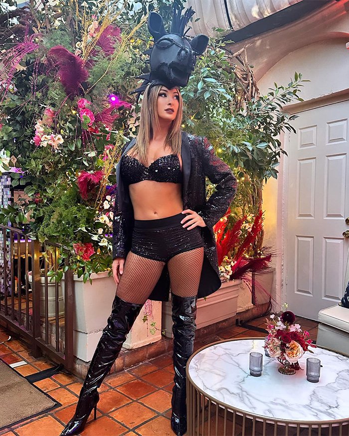 Woman dressed in black sequined outfit and tall boots posing indoors with elaborate headpiece and floral background, former adult star.