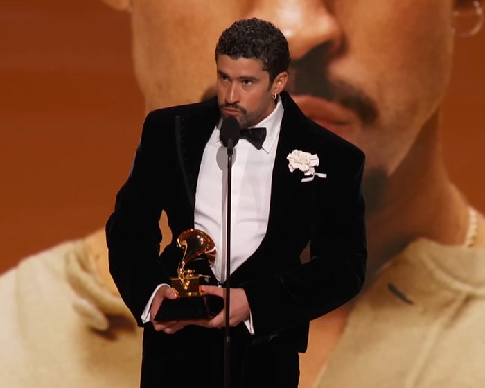 Man in tuxedo holding a Grammy award on stage amid wild theories of celebrities with $2 bills at the Grammys event.