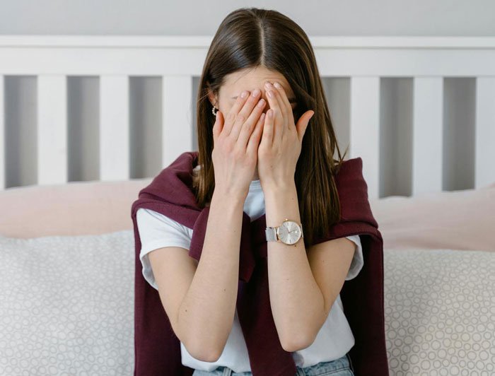 Woman covering her face with hands sitting on bed, showing distress after mom&rsquo;s joke about calling CPS on her.