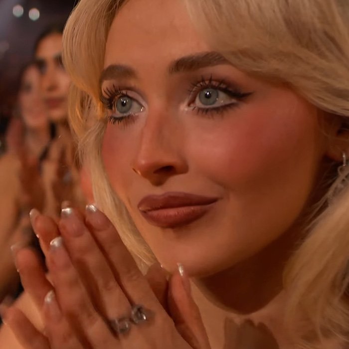 Close-up of a woman with blonde hair and blue eyes clapping during a viral claim debate about men like Sydney Sweeney and women like Sabrina Carpenter.
