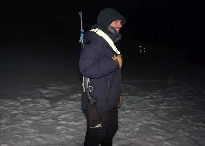 Content creator exploring the world’s darkest island at night, dressed warmly with a rifle on snowy terrain.