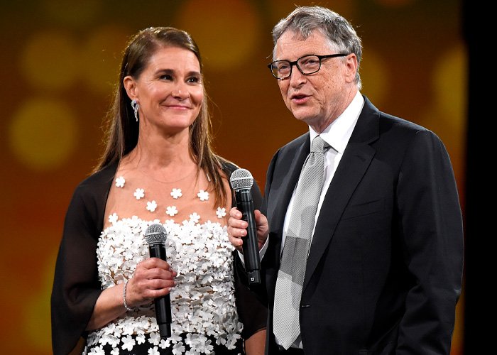 Melinda Gates and Bill Gates speaking at an event, holding microphones in a formal setting with warm lighting.