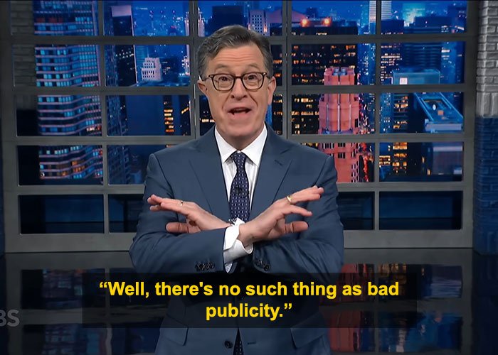 Stephen Colbert in a suit and glasses making a hand gesture on a late-night show set discussing Epstein files response.