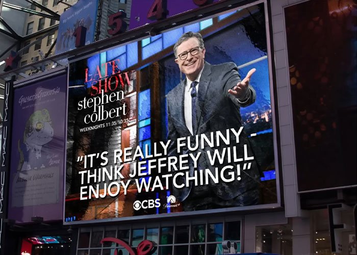 Stephen Colbert on a large billboard in Times Square, smiling and gesturing during The Late Show taping.