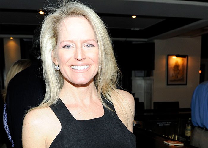 Woman with blonde hair wearing black sleeveless dress, smiling indoors with dim background, related to Stephen Colbert Epstein files viral response.