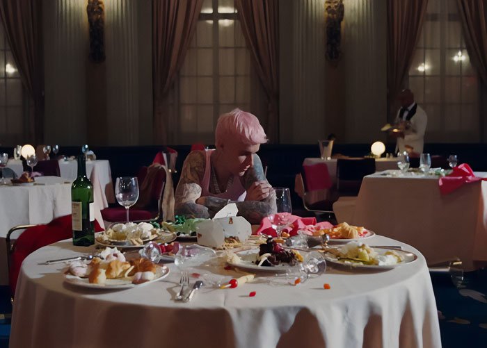 Justin Bieber sitting alone at a table with messy dishes and wine in a dimly lit room, referencing Yummy music video.