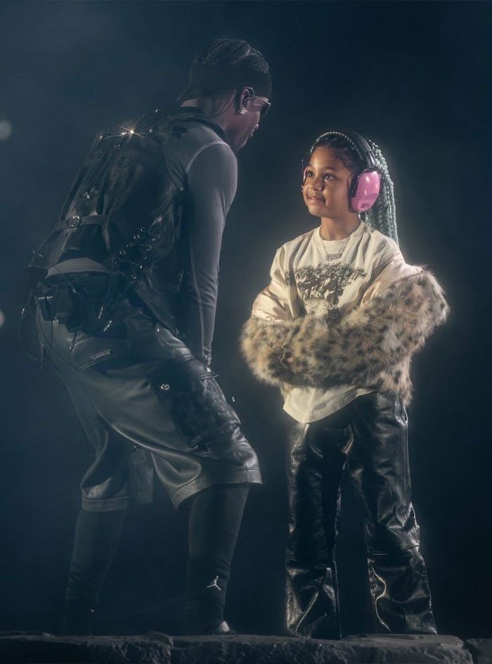 Travis Scott and child at event, wearing headphones and stylish leather outfits in dimly lit setting.